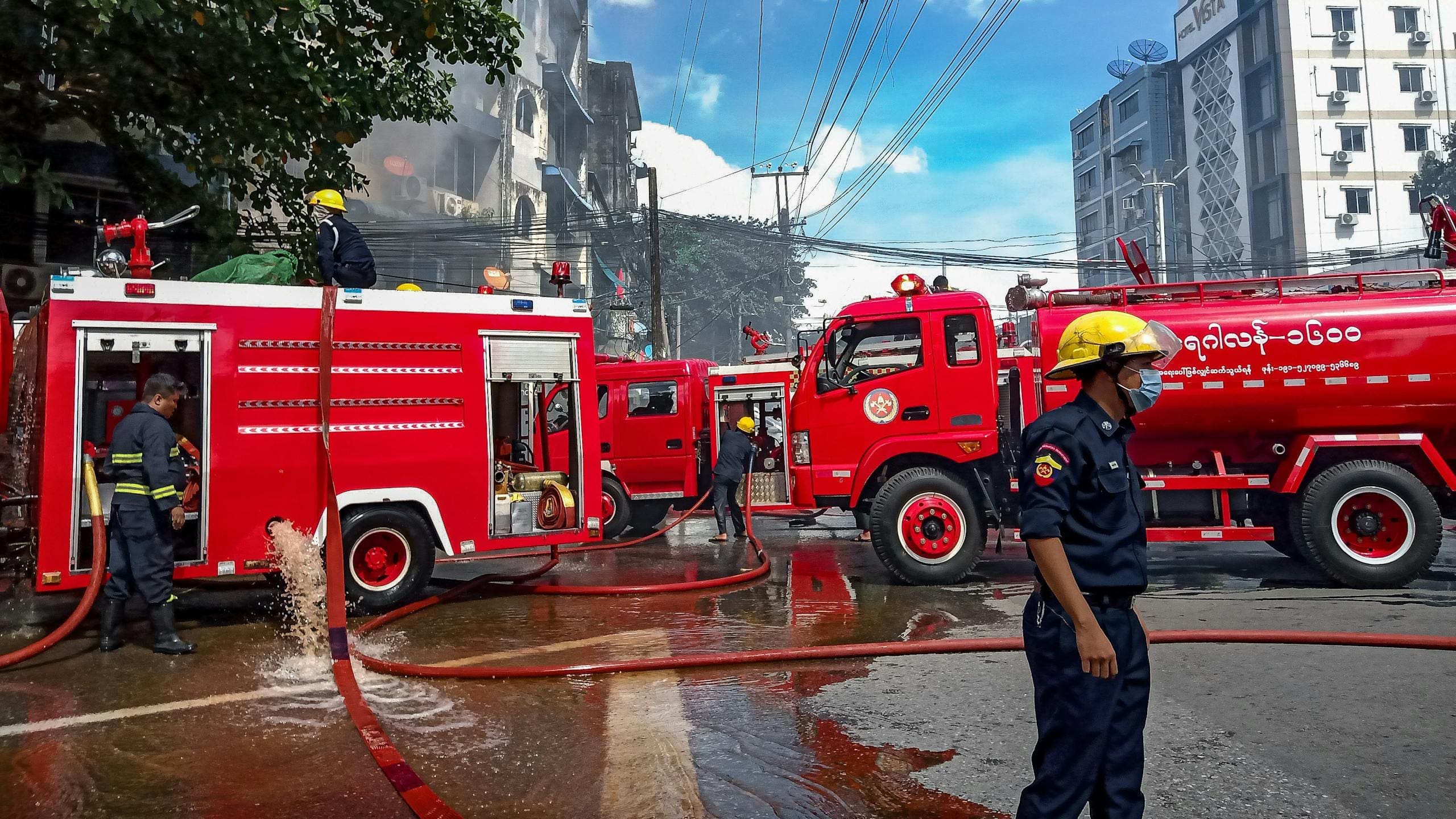 Firefighting team battling a building fire in Yangon, Myanmar with red fire trucks and hoses in action.