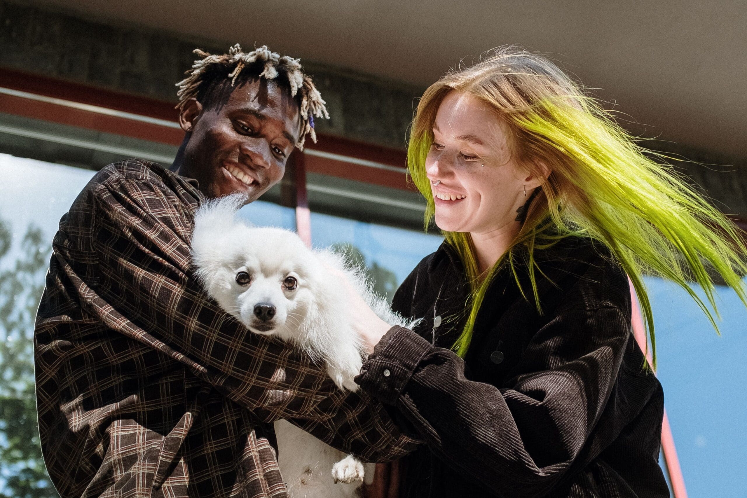 Black man with short dreadlocks and white woman with lime green-dyed hair smiling and holding a cute, white dog together.
