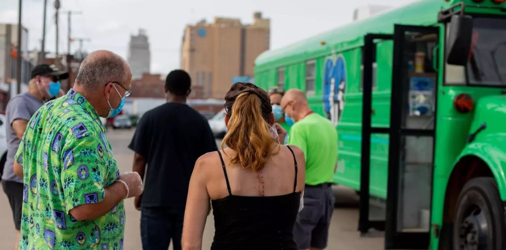 Man and woman face away from the camera outdoors with the downtown Kansas City skyline in the background. On the right is a green school bus