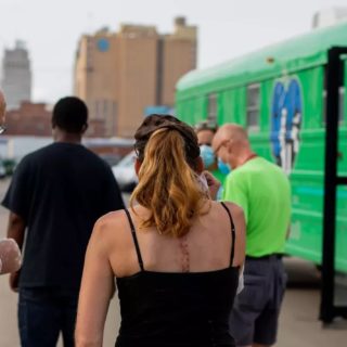 Man and woman face away from the camera outdoors with the downtown Kansas City skyline in the background. On the right is a green school bus