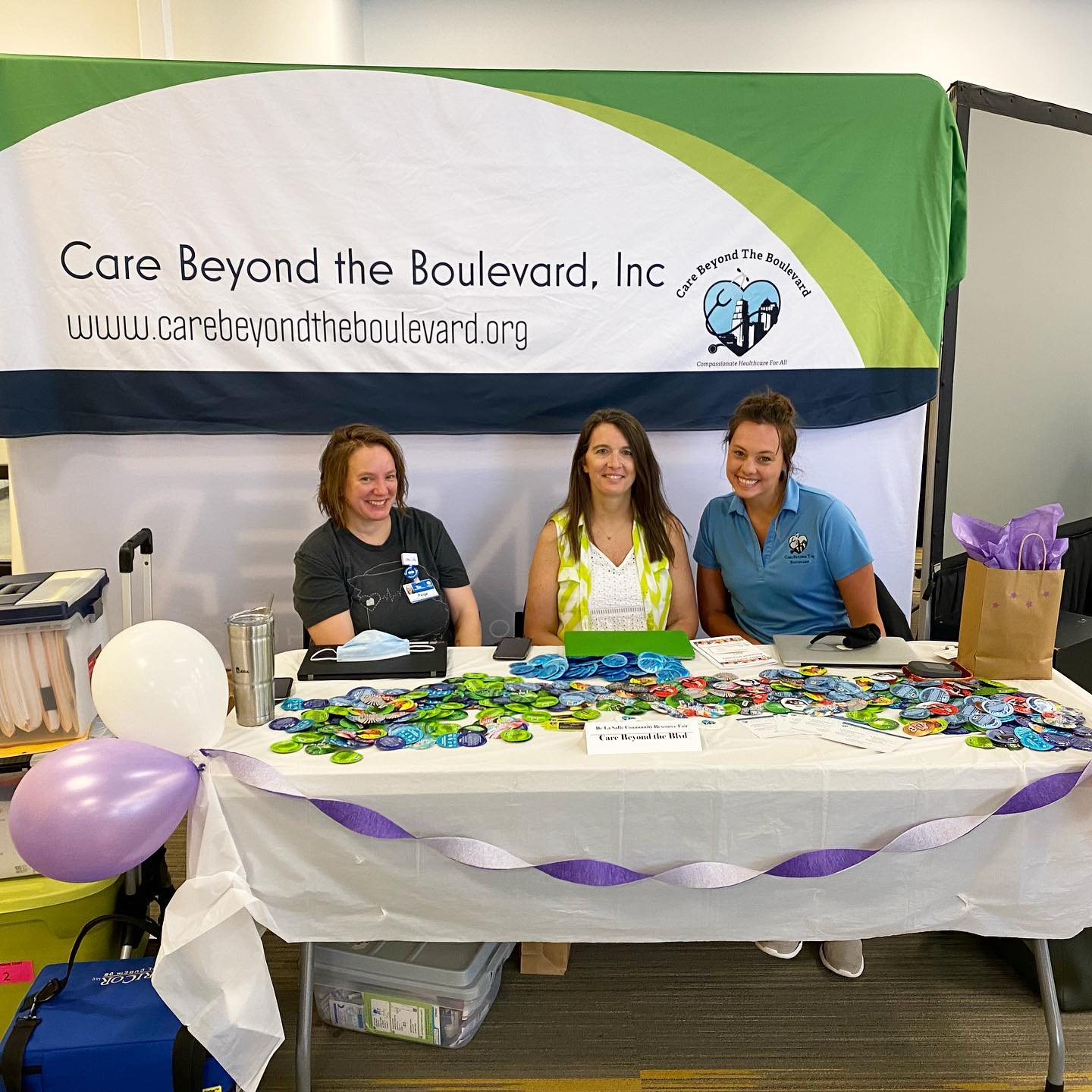 Three woman sit behind table covered in small freebies and smile at the camera. Behind them is a Care Beyond the Boulevard banner