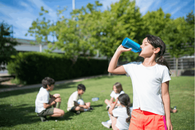 Following cool tips for a healthy summer, a girl drinks from a water bottle in front of friends who are taking a break from playing soccer outside.