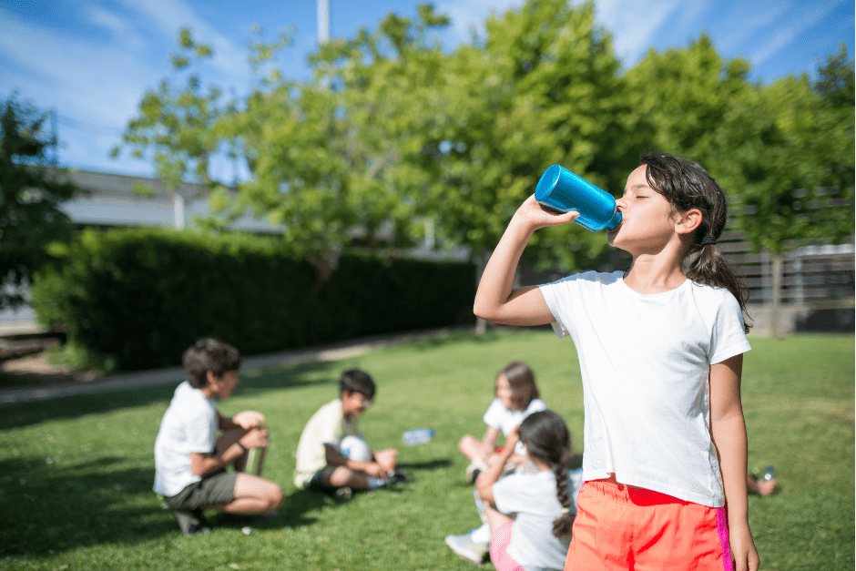 Following cool tips for a healthy summer, a girl drinks from a water bottle in front of friends who are taking a break from playing soccer outside.
