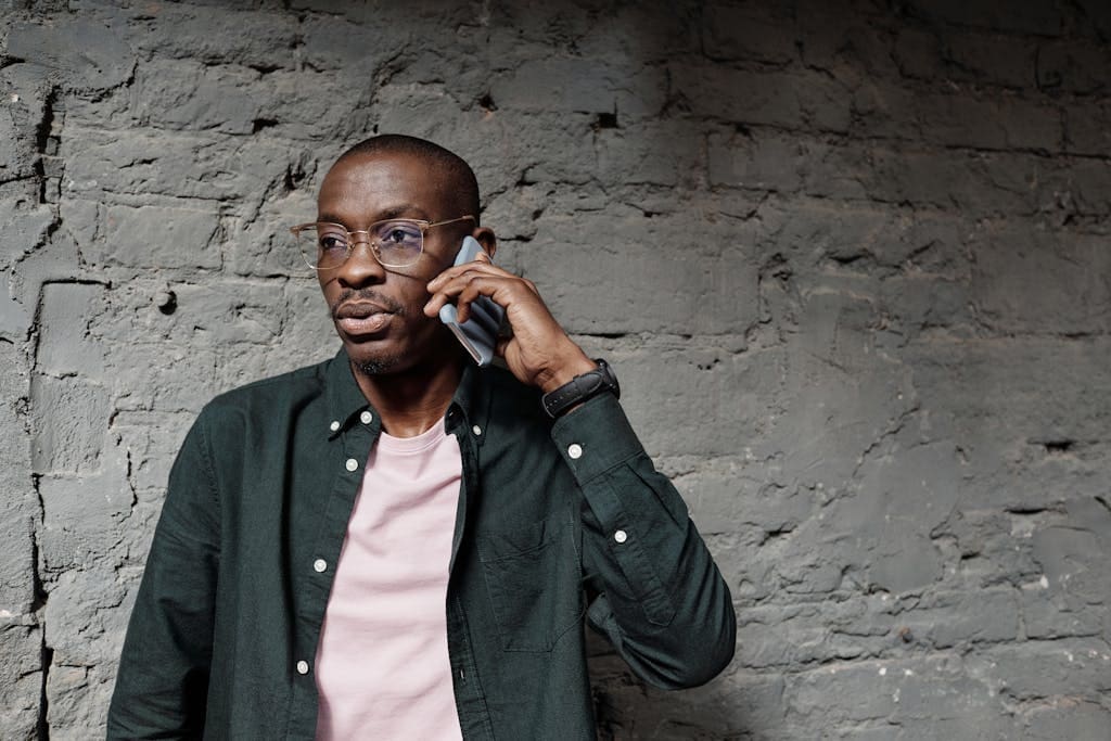 African American man in glasses talking on smartphone with casual attire against concrete wall.
