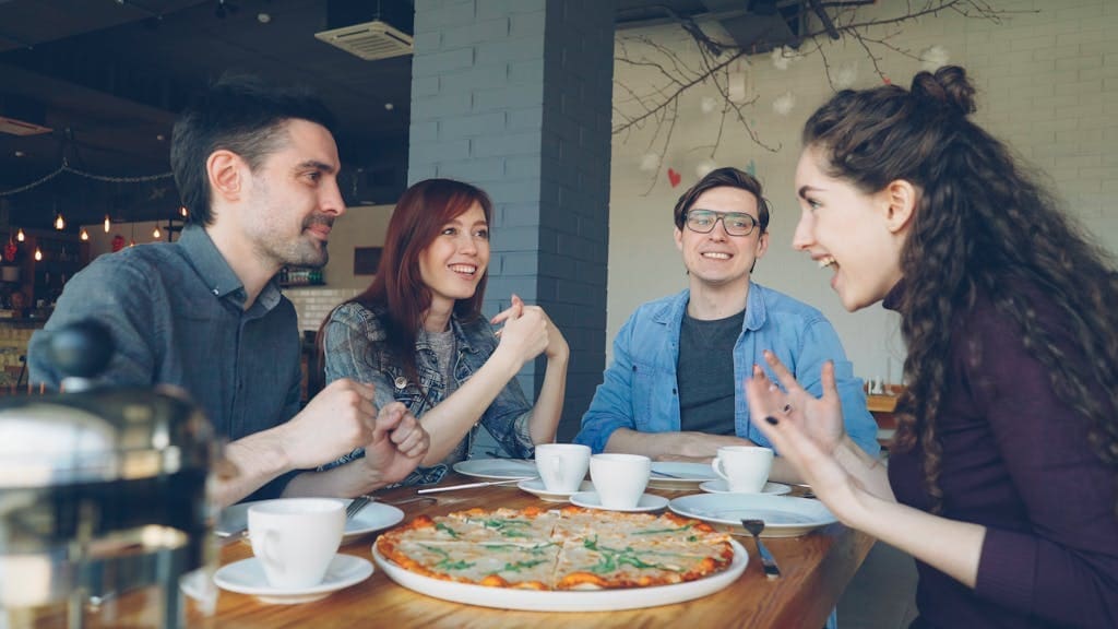 Group of four friends laughing and enjoying pizza and coffee together in a cozy cafe.