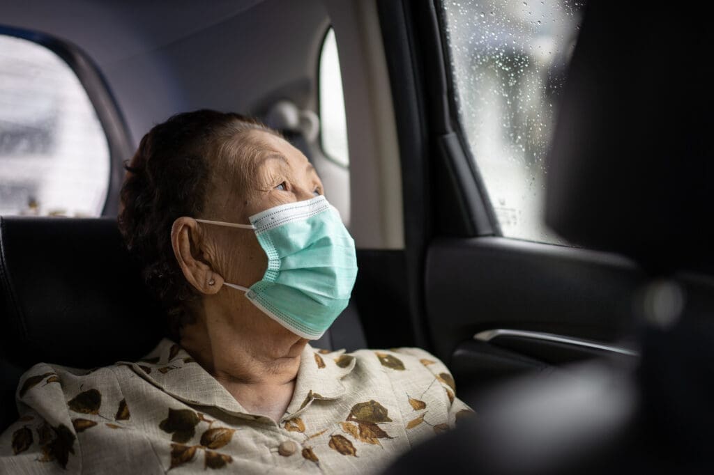 An elderly woman wears a medical mask as she rides in the back seat of a car.